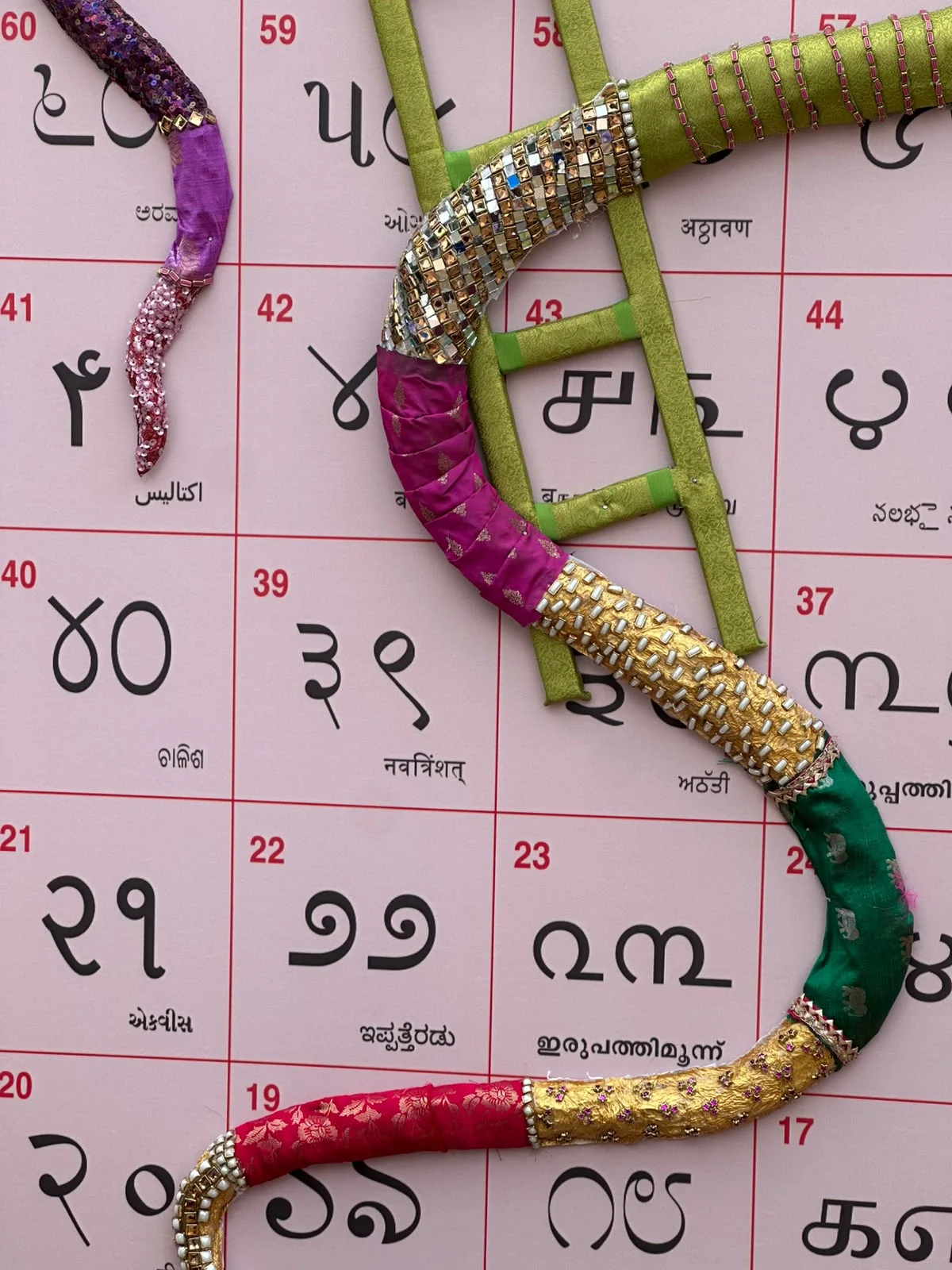 Close-up of a fabric snake wrapped in pink, green and gold brocade with sequins, winding over squares 17 to 44 next to a green ladder.