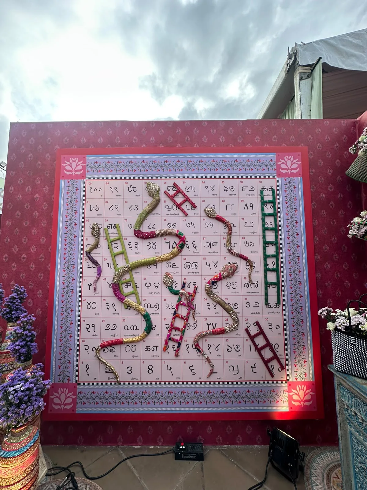 The finished snakes and ladders board mounted on a red printed wall in the PinQ booth, with fabric snakes and ladders and flower arrangements on either side.