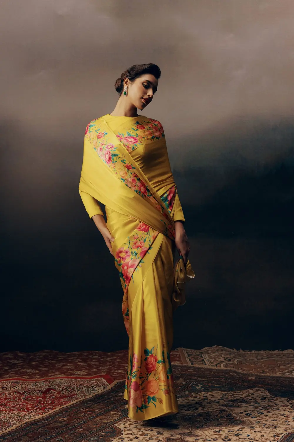 Model in a yellow floral saree on a Persian rug against a dark painted sky